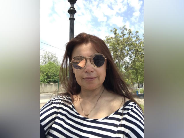 MadamXDream has brown hair, wears round sunglasses, and a black-and-white striped shirt. She's smiling slightly with a necklace. Her tits are small, and she's outside.