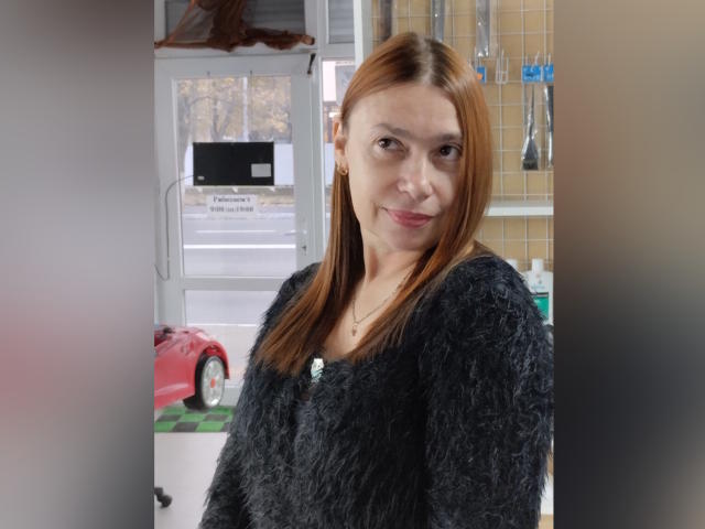 MadamXDream has straight red hair, a cute face, and small tits, wearing a fluffy black sweater, standing in a kitchen with a red car outside the window.