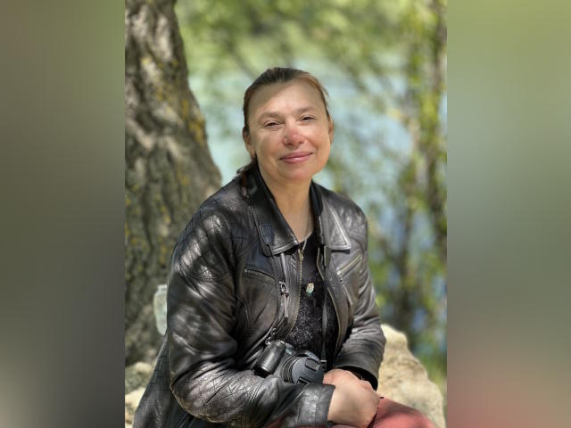 MadamXDream, with brown hair in a ponytail, smiling, wearing a shiny black leather jacket, sitting outdoors, hands on knees, green tree background, natural light, casual look.