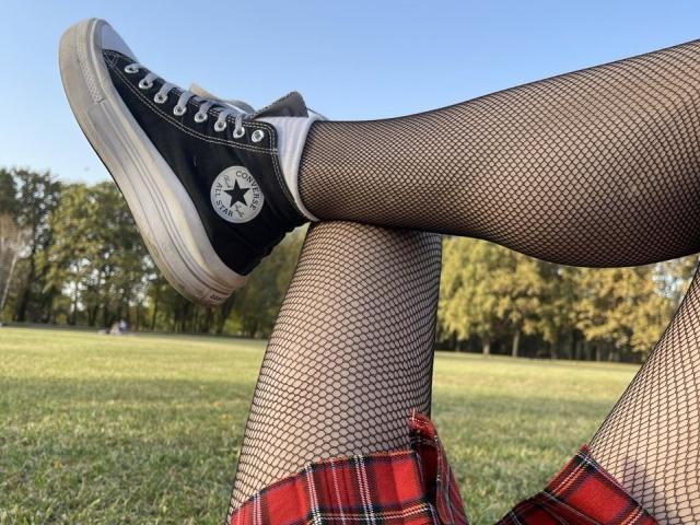 Tsukerberg's legs in fishnet stockings, black Converse sneakers with white soles, and red plaid skirt, raised in a grassy field with a clear blue sky.
