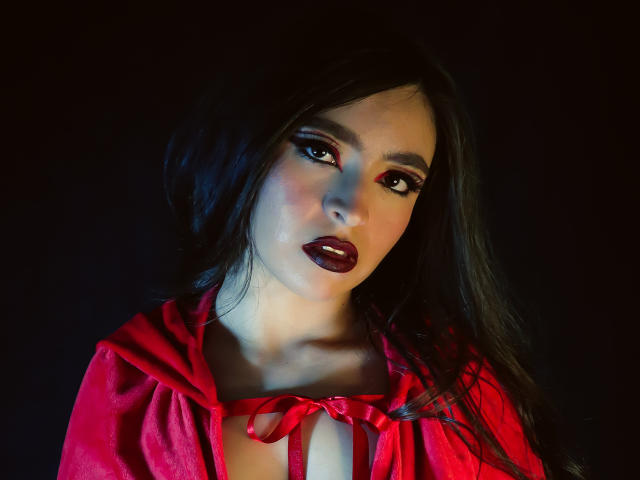 EvaMcAllister with dark hair, red lipstick, and red dress, looking seductively at the camera, her tits partially visible, red ribbon tied around her chest, dark background.