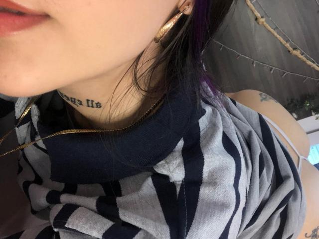 Le visage de CieloSmith, avec un tatouage sur le cou, des cheveux teints en violet, des boucles d'oreilles dorées, et portant une chemise rayée, laissant apparaître un tatouage sur l'épaule et une bretelle blanche..