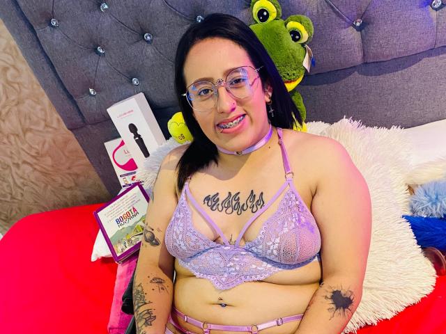 NatalieHarris with glasses, wearing lavender lace bra, showing her tits, tattoos, and belly button, smiling on a bed with a green stuffed frog. Her pussy is not visible.