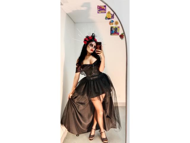 EmilyJensenn in a black, off-shoulder, corset-style dress with a sheer, flowing skirt, black high heels, and a red-black flower headpiece, holding a phone in front of a mirror.