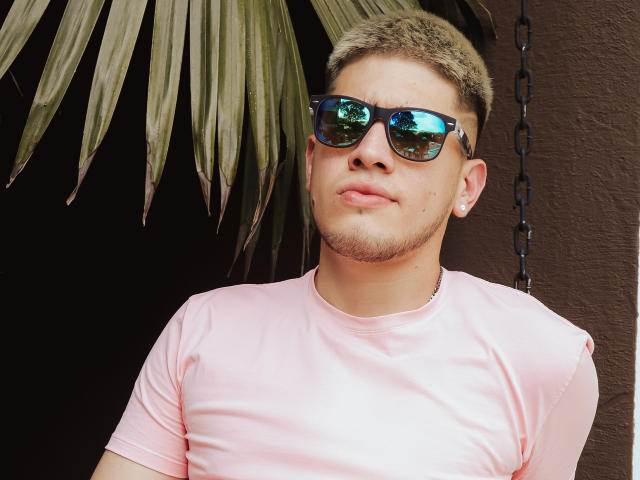 SebastiianMaxime, avec ses cheveux blonds courts, portant des lunettes de soleil bleues réfléchissantes, un t-shirt rose et des boucles d'oreilles de perles, a l'air légèrement ennuyé. Ses fesses ne sont pas visibles..