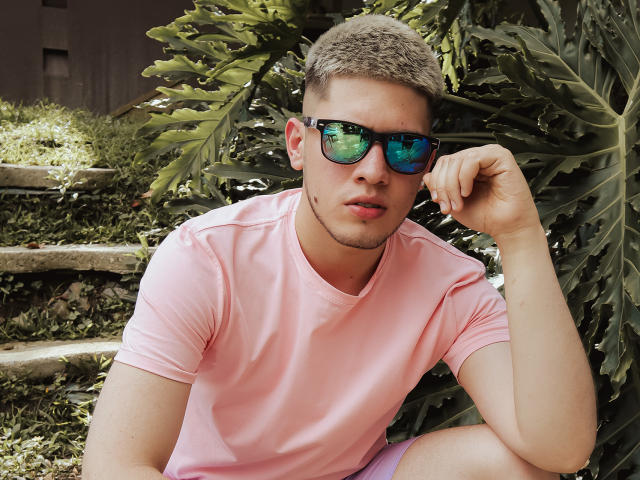 SebastiianMaxime mit kurzen, braunen Haaren, der ein pinkes T-Shirt, blau-grüne, spiegelnde Sonnenbrillen und pinke Shorts trägt, sitzt an grünen Blättern und berührt sein Kinn mit einem entspannten Ausdruck..