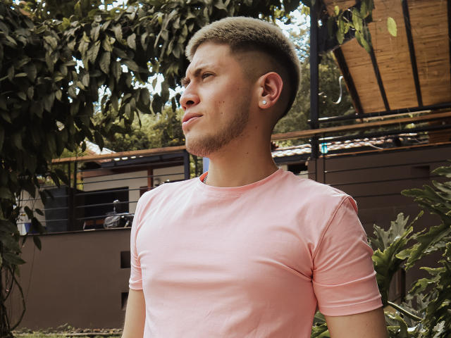 SebastiianMaxime, um cara bonitinho com cabelo raspado, vestindo uma camiseta rosa, olhando para o lado com um leve sorriso, em frente a algumas plantas..