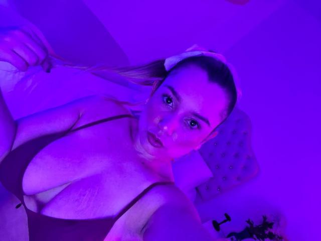 AmayaJhons has big boobs in a dark bra, purple neon lighting, and her hand is in her hair, looking at the camera with a slightly open mouth.