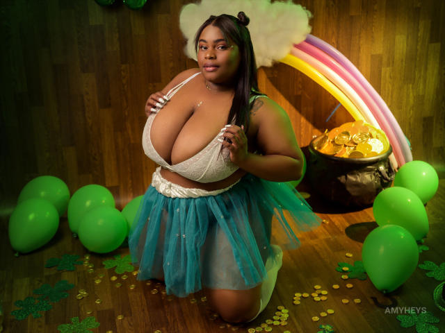 ClarieHaris with huge tits, wearing a white lace bra and blue tulle skirt, kneeling on wooden floor, surrounded by green balloons and yellow confetti, with a rainbow and pot.