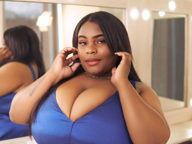 Clarie Haris, mit dunkler Haut und langen schwarzen Haaren, trägt ein blaues Kleid mit großer Brust und blickt in einen Spiegel, wobei ihre Hände in der Nähe ihrer Ohren sind. Sie lächelt leicht. Die Spiegelung ist sichtbar..