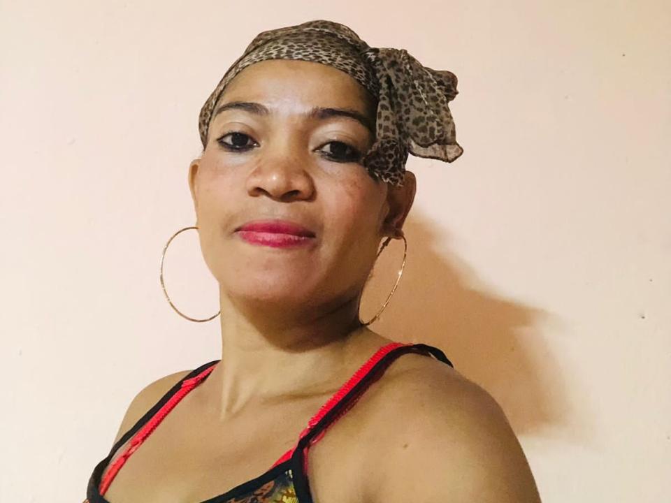 ZarahWish tiene la piel oscura, lápiz labial rosa, y lleva un pañuelo en la cabeza con estampado de leopardo, aretes de aro dorados y un top negro con tirantes rojos. Está sonriendo ligeramente..