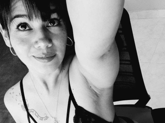 Black-and-white selfie of sashasweetx with short dark hair, hoop earrings, and a tattoo on her right shoulder, raising her arm, showing her small tits in a lace top.