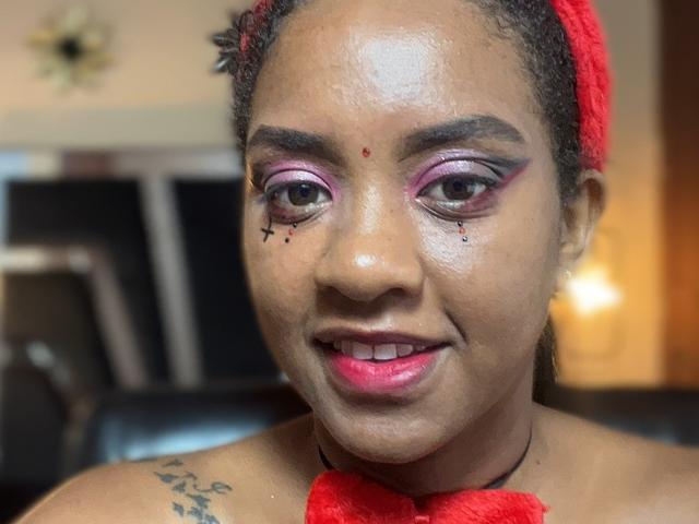 TiffanySammy with dark skin, red headband, pink eyeshadow, black eyeliner, red lipstick, small red dot on forehead, tattoos on shoulder, smiling, bare shoulders, light skin glow.