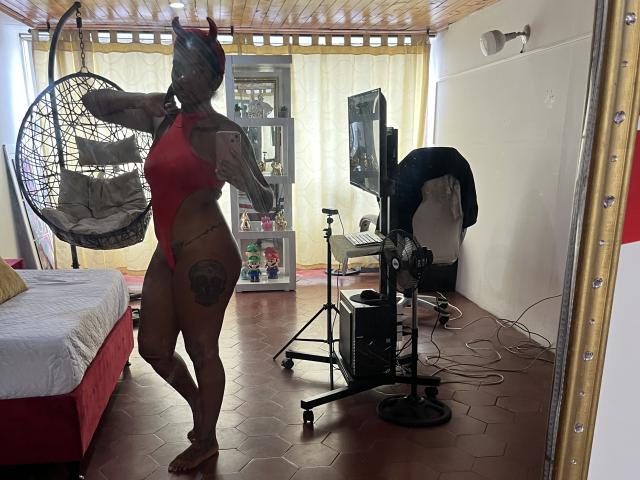 TiffanySammy, dark-skinned, muscular, in a red thong leotard, holding a hanging chair, with a large tattoo on her right thigh, standing in a room with a mirror.