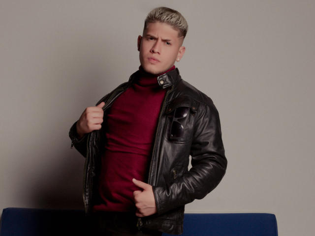SebastiianMaxime se tient debout, vêtu d'une veste en cuir noir, d'un col roulé rouge et d'un pantalon bleu, et pointe sa poitrine avec une expression sérieuse, tout en tenant le col de sa veste d'une main..