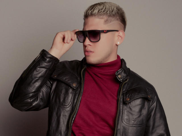 SebastiianMaxime, avec ses cheveux blonds décolorés, sa veste en cuir noir, son col roulé rouge et ses lunettes de soleil noires, ajuste ses lunettes avec une expression sérieuse, et porte une boucle d'oreille en perle..