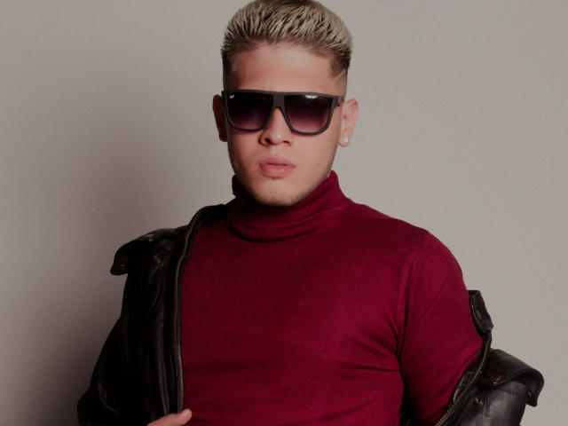 SebastiianMaxime mit blonden, stacheligen Haaren, schwarzen Sonnenbrillen, einem roten Rollkragenpullover und einer schwarzen Lederjacke, wirkt ernst, Hand auf der Jacke..