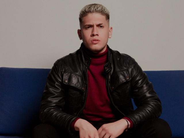 SebastiianMaxime with bleached blonde hair, serious expression, wearing a black leather jacket, red turtleneck, and black pants, sitting on a blue sofa. Hands resting on knees.