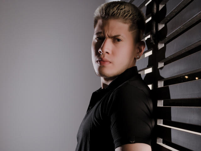 SebastiianMaxime mit ernstem Gesichtsausdruck, vor einer Wand mit Lamellen stehend, trägt ein schwarzes Hemd, blickt zur Seite, mit leicht zusammengezogenen Augenbrauen und blonden, hochgesteckten Haaren..