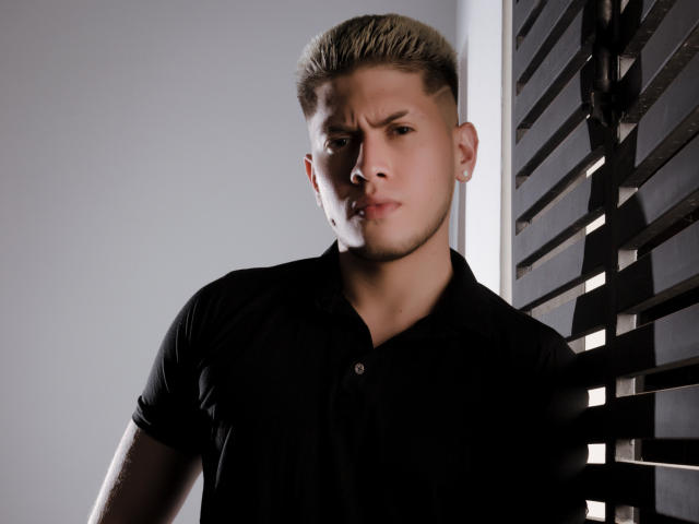 SebastiianMaxime, capelli biondi e corti, camicia nera, espressione seria, in piedi di fronte a una parete scura con listelli, luce laterale, viso leggermente in ombra, mano destra che tocca la parete..