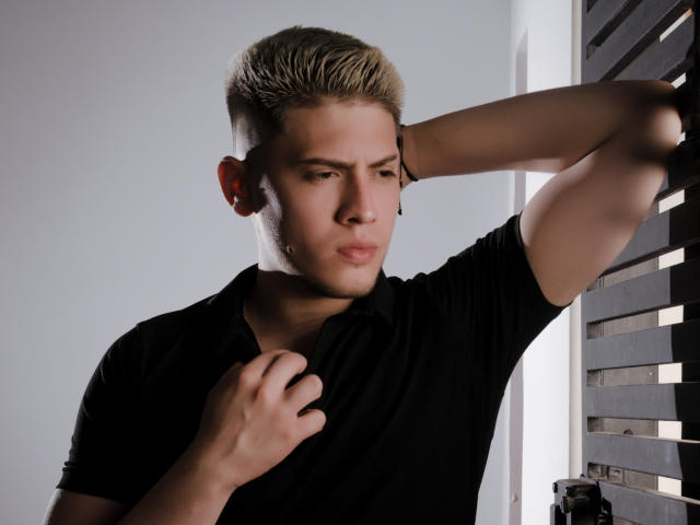 SebastiianMaxime tiene el cabello rubio y puntiagudo, viste una camiseta polo negra, con una mano en el pecho y el otro brazo levantado, con una expresión de puchero. Sus glúteos son ligeramente visibles..