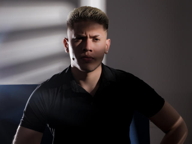 SebastiianMaxime has a serious expression, short blond hair, and a black polo shirt. He's standing with hands on hips, light shadows on his face.