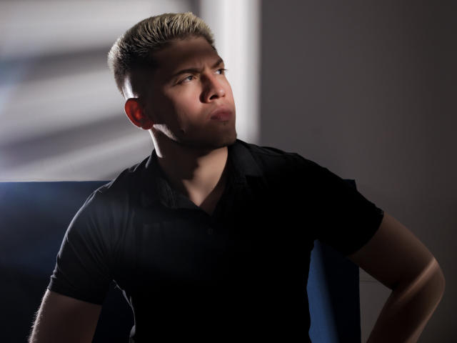 SebastiianMaxime, con i capelli biondi corti, un'espressione accigliata, una maglietta nera, braccia muscolose, che guarda di lato, in un ambiente poco illuminato, con un'ombra sul viso, una luce soffusa proveniente da sinistra..