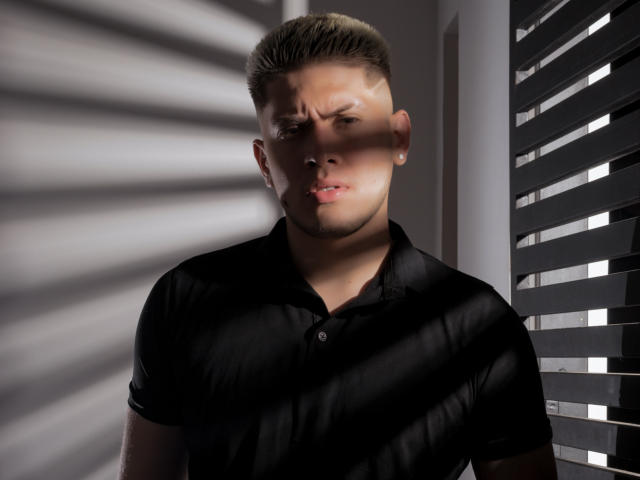 SebastiianMaxime, con capelli biondi corti, una polo nera, un'espressione seria, la luce del sole che proietta ombre sul suo viso, in piedi vicino a delle persiane nere, con una leggera barba di tre giorni, un piccolo piercing all'orecchio, corporatura muscolosa..