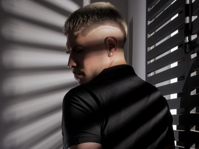 SebastiianMaxime con un peinado estilo mohawk, perfil lateral, camisa negra, perforación en la oreja, con sombras proyectadas por persianas, mirando hacia abajo, contraste de luz y sombra, cabello corto, rasgos faciales sutiles, expresión seria..