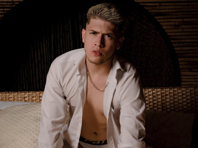 SebastiianMaxime, con i capelli biondi, corti e dall'aspetto bagnato, una camicia bianca aperta che lascia intravedere il petto nudo e una catena d'oro, è in avanti con un'espressione seria e leggermente infastidita..
