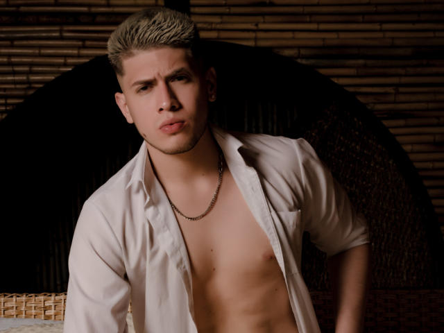 SebastiianMaxime with blonde hair, white shirt open, showing abs, wearing a chain necklace, looking annoyed, standing in front of a wicker chair.