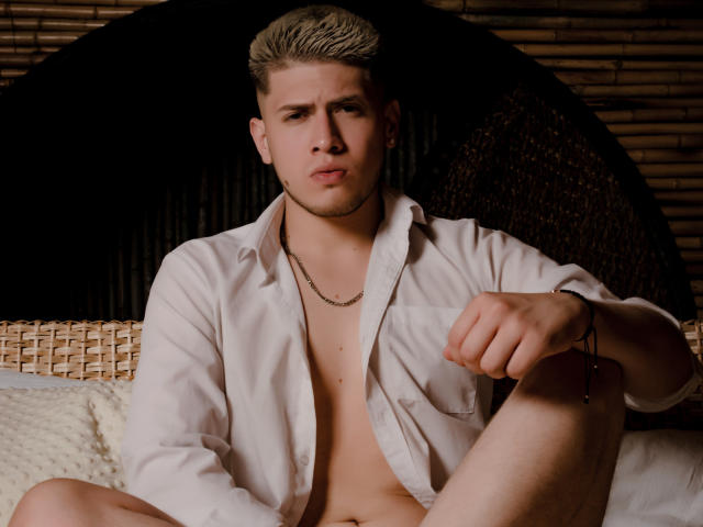 SebastiianMaxime, rubio, camisa blanca, sin pantalones, trasero expuesto, sentado, silla de mimbre, fondo de madera, expresión seria, collar, pulsera, camisa ligeramente abierta, brazo derecho apoyado en la rodilla..