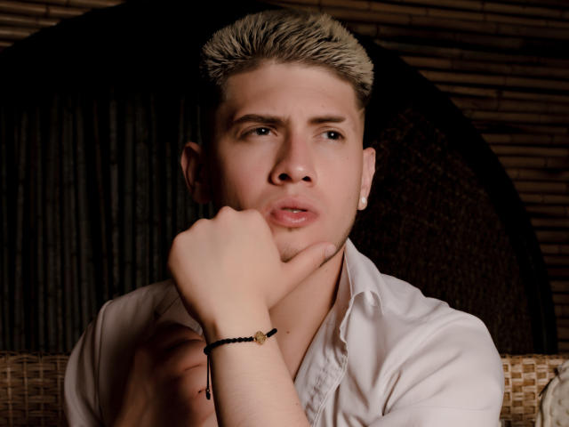 SebastiianMaxime, avec ses cheveux blonds hérissés, sa chemise blanche et son bracelet noir, a une main sur son menton, l'air légèrement confus, avec une peau pâle et de petites boucles d'oreilles blanches..