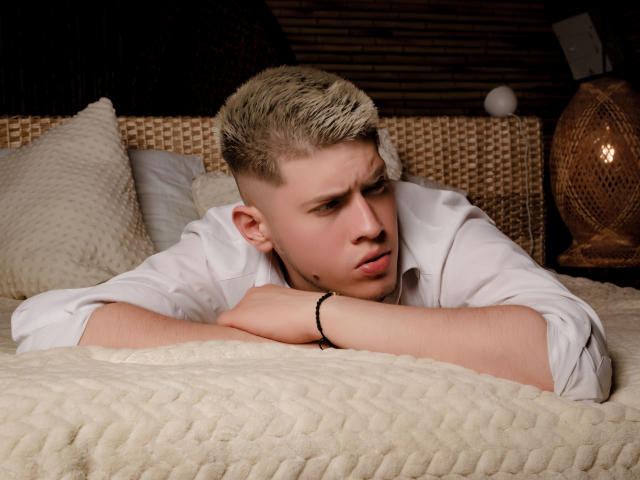 SebastiianMaxime with blond buzzcut, white shirt, black bracelet, lying on bed, arms crossed, looking annoyed, butt up, wavy white blanket, wicker headboard, lamp in background.