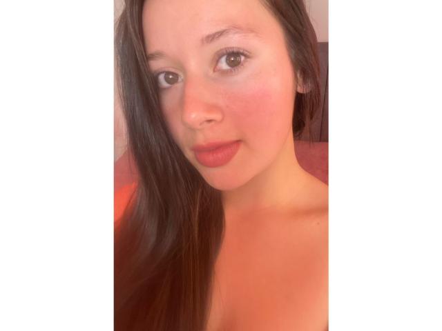 DaphneEvans69 tem pele clara, olhos castanhos, cabelos castanhos longos, lábios rosados e um rosto levemente avermelhado. Seus ombros estão à mostra. Ela olha diretamente para a câmera..