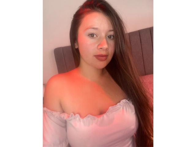 DaphneEvans69, com cabelos castanhos longos, rosto com tons avermelhados, vestindo uma blusa branca com um ombro descoberto, mostrando seus seios, e com uma expressão séria, sentada encostada em uma cabeceira cinza..