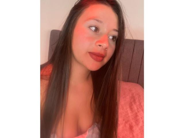 DaphneEvans69, con lunghi capelli castani, rossetto rosso e un carino piercing al naso, mostra il seno indossando una maglietta rosa, guardando di lato con un leggero sorriso..