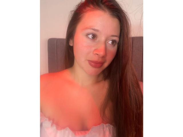 Daphne Evans, 69 ans, avec de longs cheveux bruns, un haut rose à une épaule, et un éclairage rouge sur son visage et sa poitrine, affichant un léger sourire et regardant sur le côté..