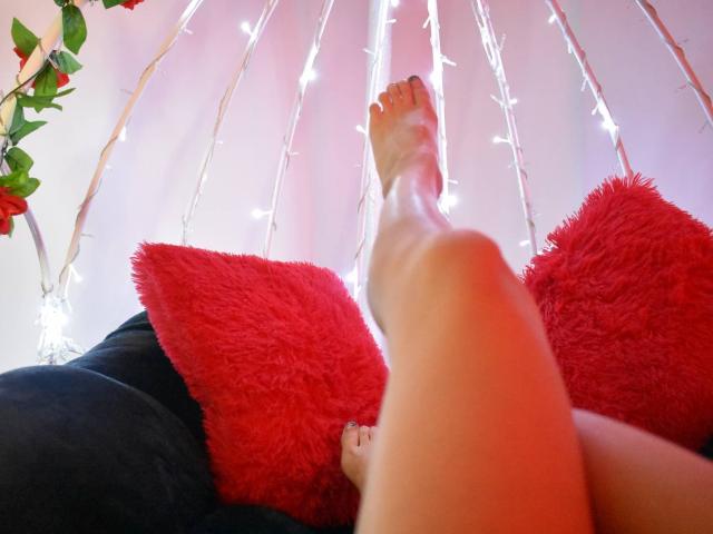 Kathrin Greys Fuß, leicht angehoben, ist im Fokus, wobei ihre Zehen nach oben zeigen. Sie sitzt auf einer schwarzen Couch mit zwei roten, flauschigen Kissen. Im Hintergrund sind Lichterketten angebracht..