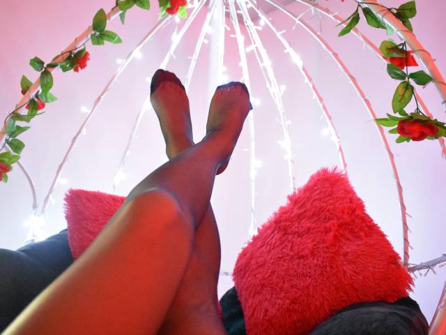 KatherinGrey's legs in black sheer stockings, crossed at the ankles, with red fluffy pillows in the foreground, white string lights, and red rose vines above.