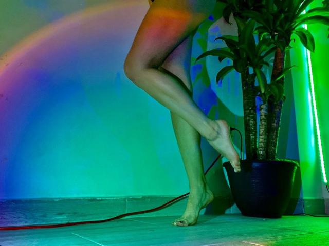 KatherinGrey's legs are crossed, showing her bare ass and feet. She's standing next to a potted plant with green and blue neon lights creating a rainbow effect.