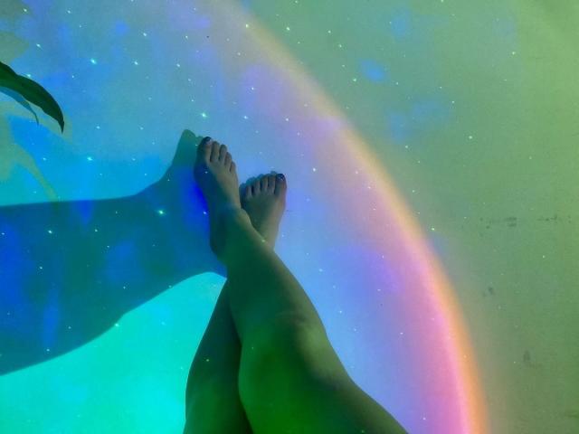 I piedi incrociati di KatherinGrey emanavano una luce arcobaleno e blu-verde, con piccole luci scintillanti, creando un'atmosfera magica e surreale. Le sue dita dei piedi erano leggermente incurvate, aggiungendo un'aria rilassata..
