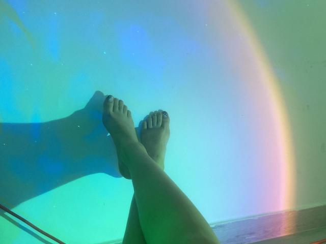 Los pies de KatherinGrey, con las uñas de los dedos pintadas de negro, están cruzados a la altura de los tobillos, sobre un degradado de verde y azul con un reflejo de arcoíris..