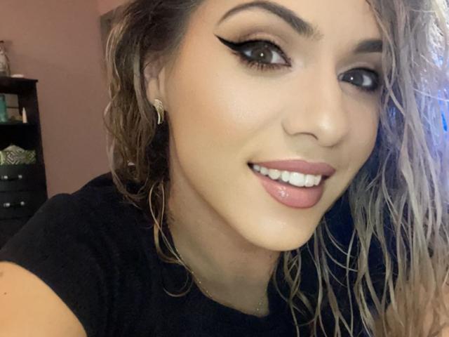 LucilleXSugar heeft golvend blond haar, donkere eyeliner, roze lippenstift, witte tanden, oorringen en een zwart shirt, en ze glimlacht naar de camera met een sexy, schattig gezicht..