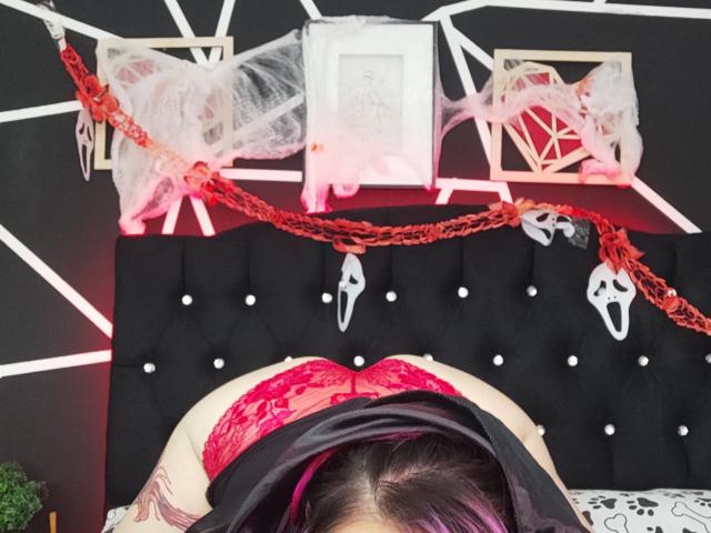 NatashaLau is bent over, showing red lace panties and a tattoo on her thigh. Black headboard with white buttons, red chains, and white masks are in the background.
