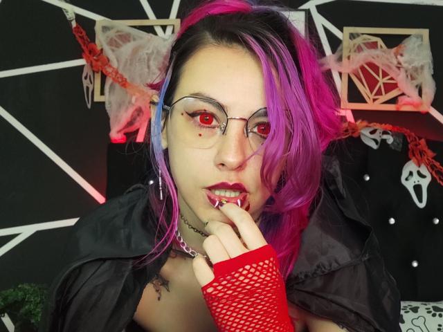 NatashaLau com cabelo roxo-rosa, luva de malha vermelha, jaqueta preta, óculos, batom vermelho, dedo na boca, olhos vermelhos, colar, fundo preto, pentagrama, decoração vermelha e branca..
