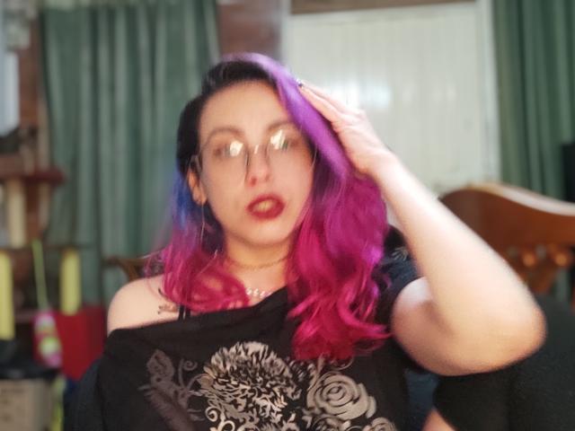 NatashaLau with purple and blue hair, wearing glasses and a black off-shoulder shirt, has her hand in her hair, showing her tits and a slight open-mouthed expression.