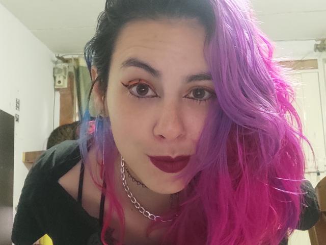 NatashaLau, con i capelli viola-rosa, rossetto scuro e eyeliner nero, guarda la fotocamera con un leggero sbuffo, indossando una maglietta nera e una collana a catena d'argento..