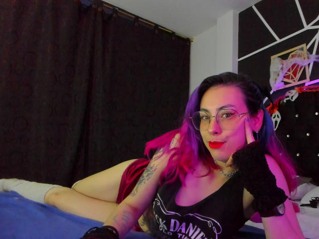 NatashaLau with purple and pink hair, glasses, black tank top, fingerless glove, tattooed arm, laying on bed, red lips, black curtains, purple and pink lighting, looking at camera.