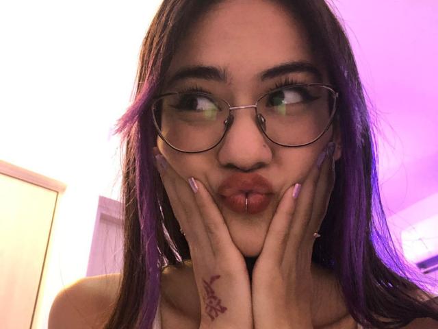 CieloSmith, avec ses cheveux violets, ses lunettes et son piercing labial, faisant la moue et posant ses mains sur ses joues, montrant un tatouage au henné sur sa main gauche, sous un éclairage rosâtre..